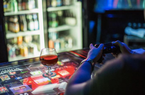 A person holds a game controller while a drink sits on a table, with a fridge full of beverages in the background.
