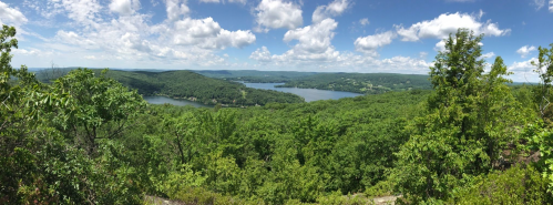 Panoramic view of lush green hills and a serene lake under a bright blue sky with fluffy white clouds.