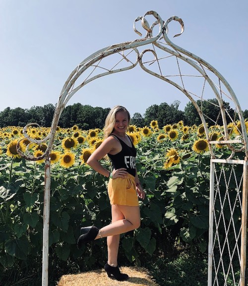 A woman poses under a decorative arch in a sunflower field, wearing a tank top and yellow shorts, smiling at the camera.