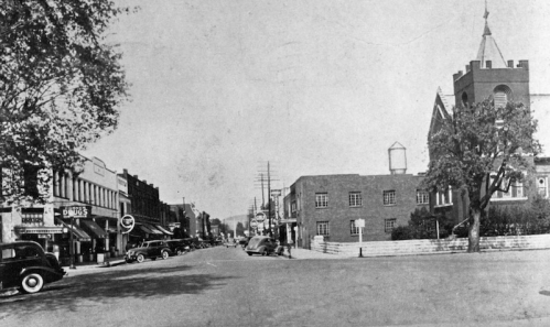 Historic black-and-white photo of a small town street with vintage cars, storefronts, and a church in the background.