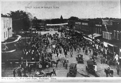 A historic street scene in Ripley, Tennessee, featuring a parade, crowds, and early 20th-century vehicles.