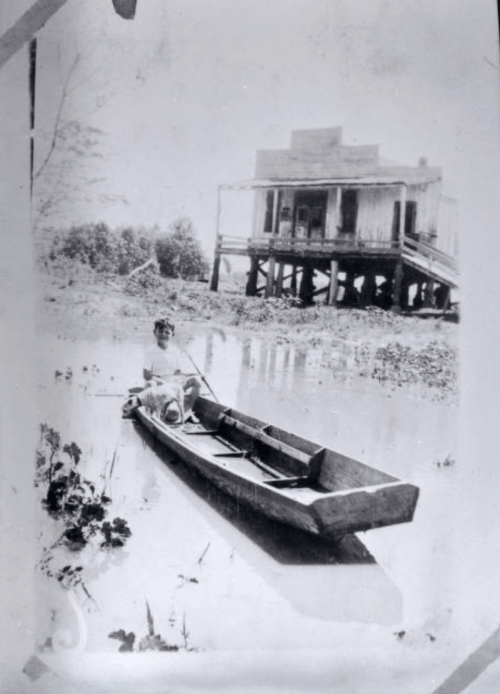 A person sits in a small boat on still water, with a raised house in the background surrounded by greenery.
