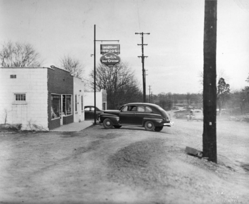 A vintage car approaches a small ice cream shop on a dirt road, with trees and power lines in the background.