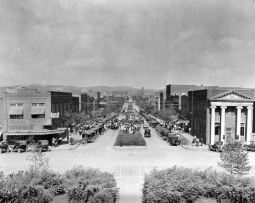 Historic black-and-white photo of a bustling street with vintage cars and crowds, lined by buildings and trees.