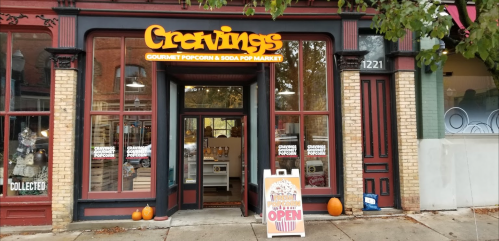 Exterior of "Cravings" gourmet popcorn and soda pop market, featuring a welcoming sign and pumpkins by the entrance.