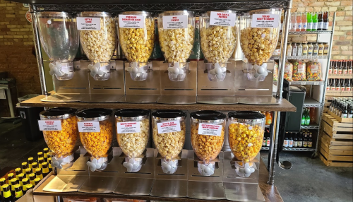 A display of popcorn dispensers with various flavors, labeled and arranged in two rows on a wooden shelf.