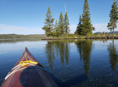 A kayak on calm water, reflecting trees and a clear blue sky in a serene natural setting.