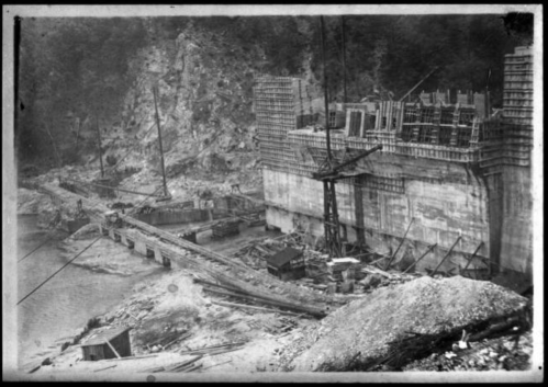 Historic black-and-white photo of a construction site with cranes and scaffolding near a riverbank.