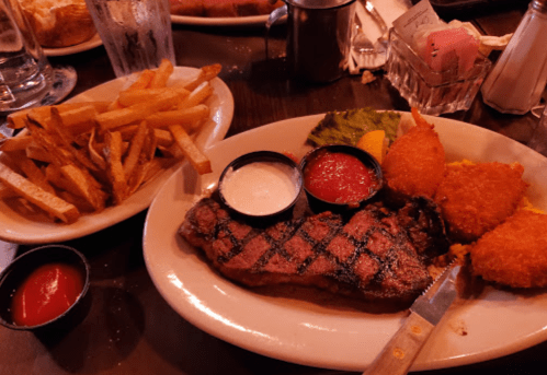 A plate with grilled steak, two dipping sauces, fried corn fritters, and a side of crispy French fries.