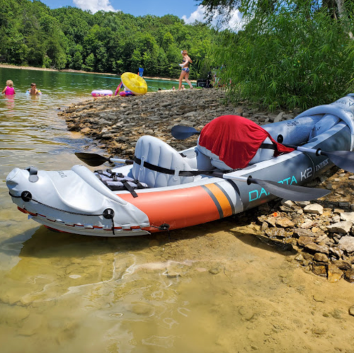 A kayak on the shore of a river, with people swimming and relaxing in the background under a sunny sky.