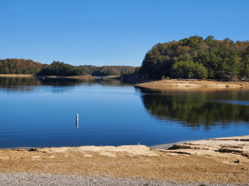 A calm lake surrounded by trees, reflecting the clear blue sky and autumn foliage on the shore.