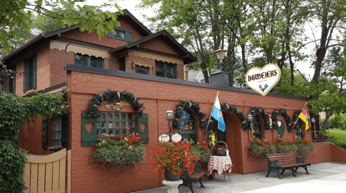 A charming restaurant with a brick exterior, green shutters, and festive decorations, surrounded by greenery and flowers.