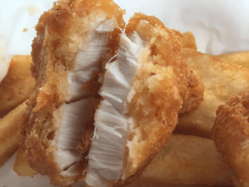 Close-up of a crispy fried chicken piece, revealing tender white meat inside, served with golden fries.