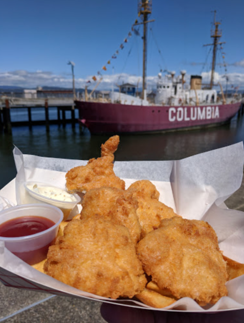 A hand holds a basket of golden fried fish with dipping sauces, with a boat named "COLUMBIA" in the background.