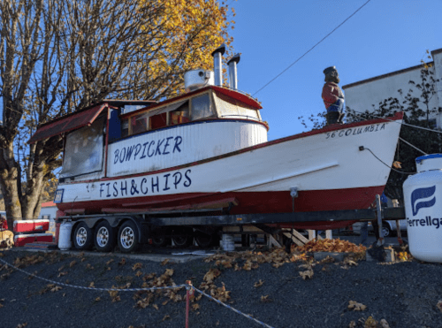 A colorful boat named "Bowpicker" with a fish and chips sign, parked on a trailer surrounded by autumn leaves.