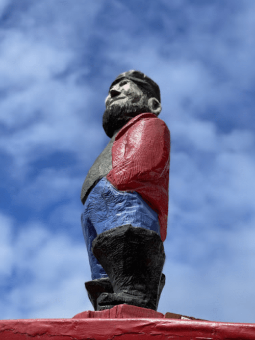 A colorful wooden statue of a man with a beard, wearing a red shirt and blue pants, against a blue sky.