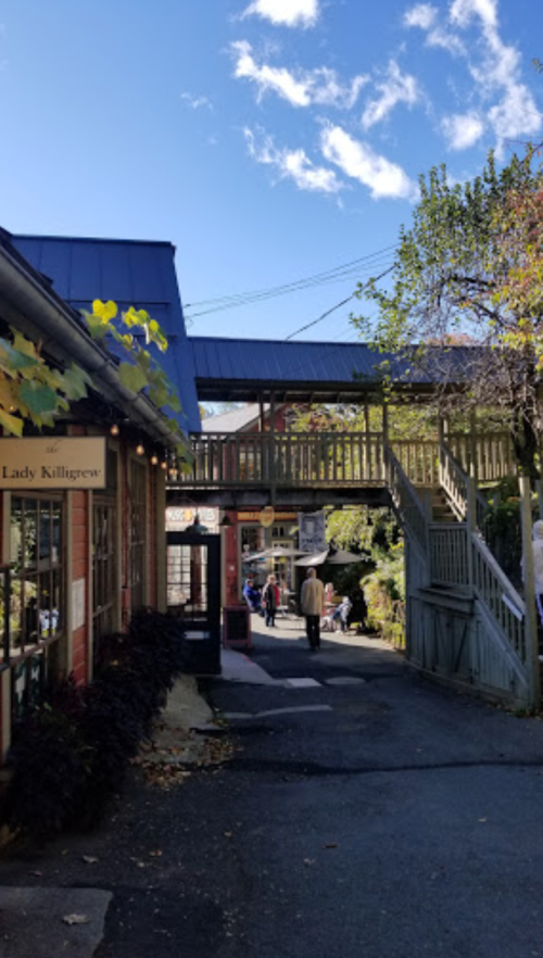 A quaint street scene with shops on either side and a bridge overhead, under a clear blue sky with scattered clouds.
