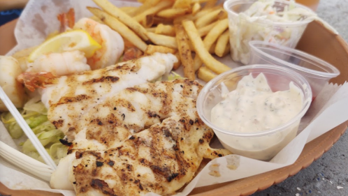 A plate of grilled fish fillets with fries, shrimp, and two cups of dipping sauce on a bed of lettuce.