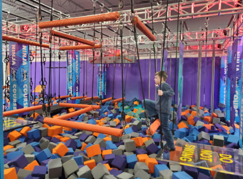 A child navigates a colorful foam pit under a suspended obstacle course in a playful indoor setting.