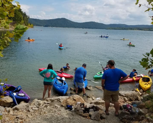 People kayaking on a lake with mountains in the background, some preparing kayaks on the shore.