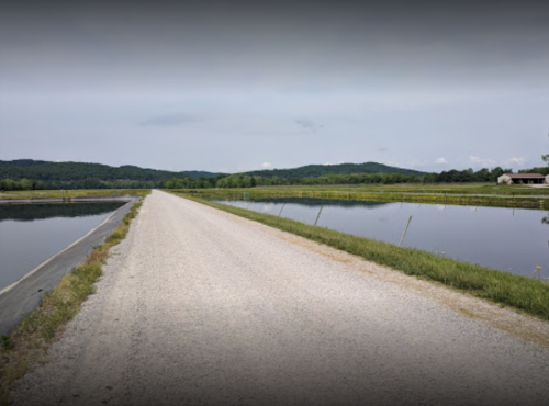 Gravel road beside calm water bodies, surrounded by green hills and fields under a cloudy sky.