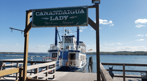 A boat named "Canandaigua Lady" docked at a wooden pier on a calm lake under a clear blue sky.