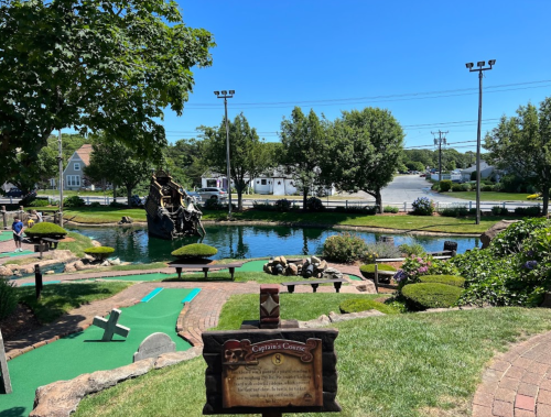 A sunny mini-golf course with a pond, trees, and a pirate ship structure in the background.