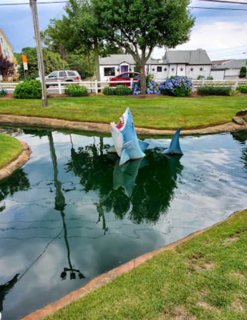 A large, cartoonish shark sculpture emerges from a pond in a mini-golf course, surrounded by green grass and trees.