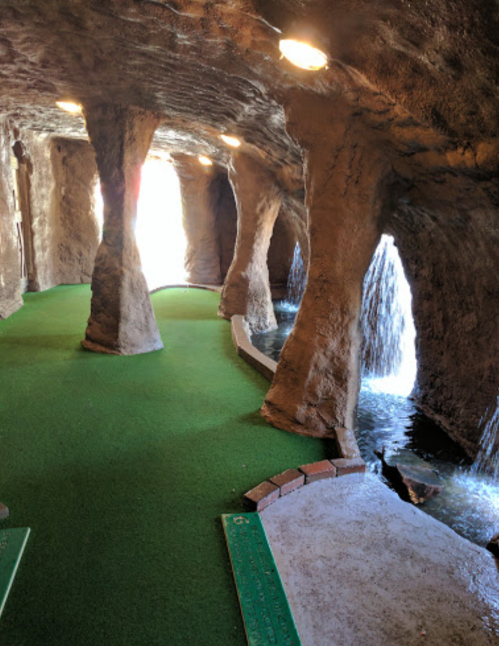 A cave-like mini-golf course with green turf, stone pillars, and a waterfall feature in the background.