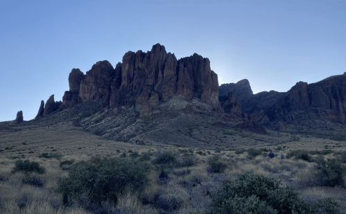 A rugged mountain range rises against a clear blue sky, with hikers visible in the foreground among desert vegetation.