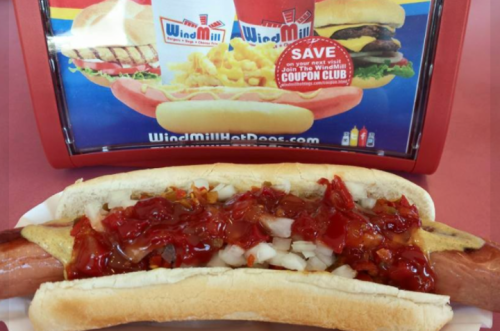 A hot dog in a bun topped with onions and ketchup, in front of a colorful menu display featuring burgers and fries.
