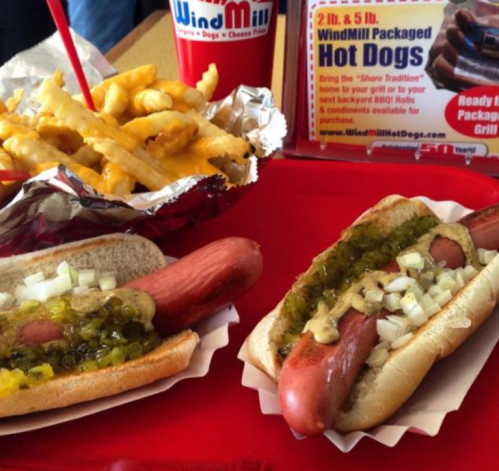 Two hot dogs with mustard, relish, and onions on a tray, accompanied by a cup of fries and a promotional sign.