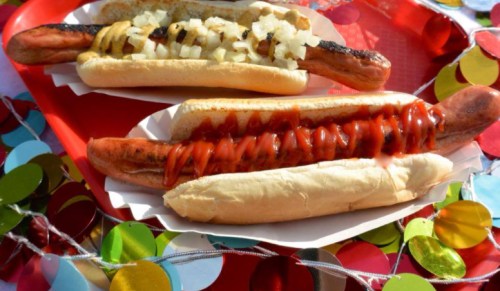 Two hot dogs on a red tray: one topped with onions and the other with ketchup, surrounded by colorful confetti.