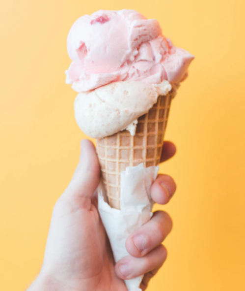 A hand holding a waffle cone with two scoops of pink and white ice cream against a bright yellow background.