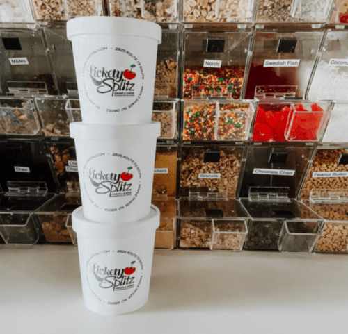 Three stacked white cups from Lickety Splitz in front of a colorful display of candy and toppings in clear containers.