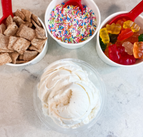 A bowl of vanilla ice cream surrounded by containers of cereal, colorful sprinkles, and gummy candies.