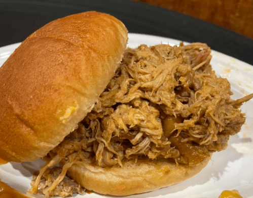 A close-up of a pulled pork sandwich on a white plate, featuring tender, shredded meat piled high in a soft bun.