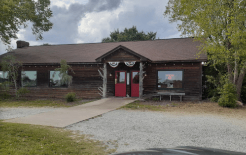 A rustic wooden building with red doors, surrounded by greenery and gravel, under a cloudy sky.