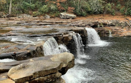 A serene waterfall cascading over rocky ledges into a calm river, surrounded by lush greenery and autumn foliage.