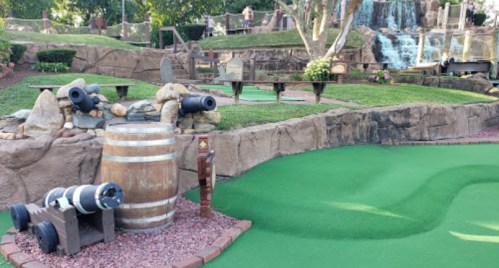 A mini-golf course featuring a wooden barrel, cannons, and a waterfall in a lush green setting.