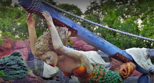 A colorful mermaid statue suspended from a wooden beam, surrounded by greenery and rocky terrain.