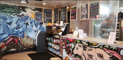 A colorful café interior featuring a graffiti wall, a menu board, and a display of snacks at the counter.