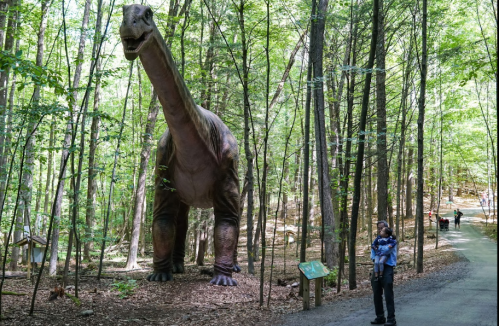 A person holds a child while standing near a large dinosaur statue in a wooded area with a path.