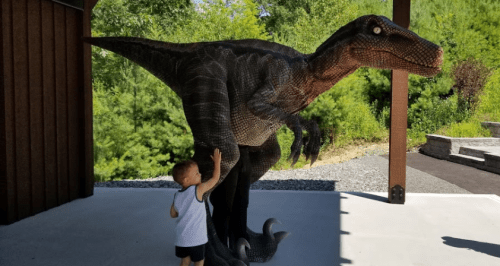 A child touches a life-sized dinosaur statue in a natural outdoor setting.
