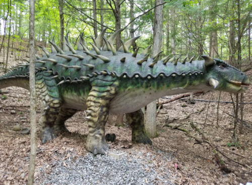 A large, spiky dinosaur statue stands among trees in a wooded area, surrounded by leaves and gravel.