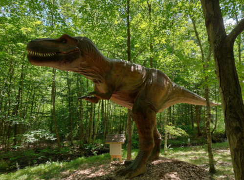 A life-sized dinosaur statue in a lush green forest, surrounded by trees and natural foliage.