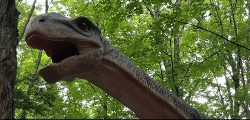 A large dinosaur model with a long neck, set against a backdrop of green trees and foliage.