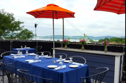A rooftop dining area with blue tables, white napkins, and orange umbrellas, overlooking a scenic view of water and hills.