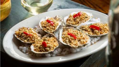 A platter of baked oysters topped with breadcrumbs and red chili, served on a bed of salt.
