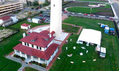 Aerial view of a lighthouse beside a historic building and a tent set up on a green lawn, with cars in the background.